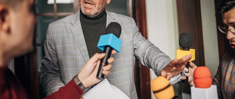 Group of people, male politician confronted by journalists with microphones.