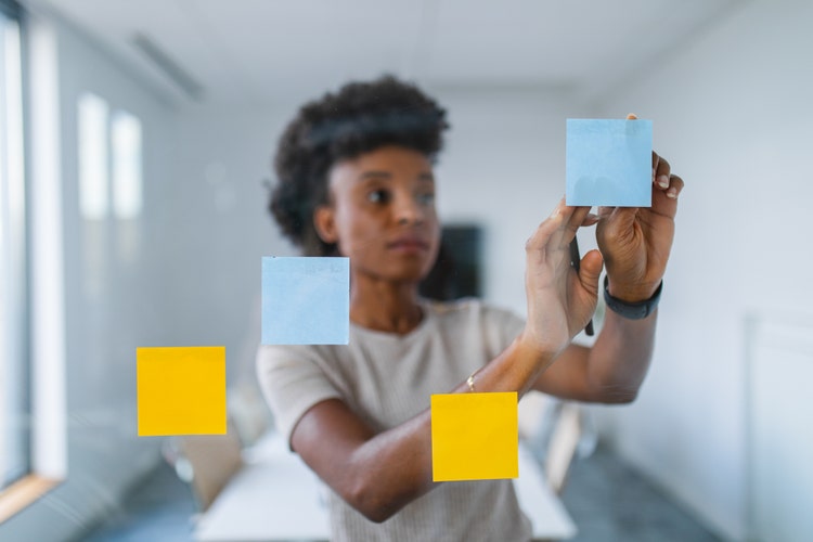 African ethnicity businesswoman engaged in creative brainstorming, placing colorful sticky notes on a glass wall in a bright office setting.