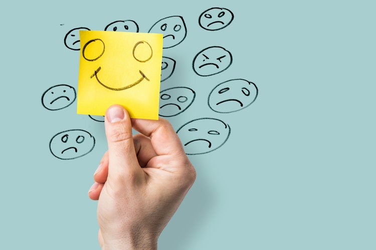 hand holding yellow notepaper showing a happy face over unhappy emoticons drawn on blue background