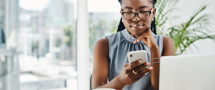 Young black businesswoman using a cellphone while working on a laptop in an office alone