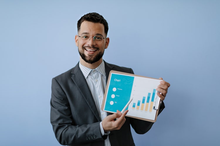 A smiling man with glasses and a beard in a suit holds up a clipboard displaying a colorful chart, standing against a light blue background in a modern office setting.