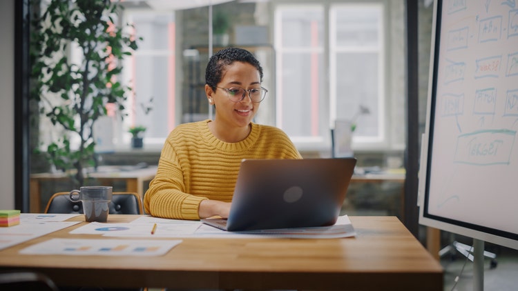 Portrait of Young Latin Marketing Specialist in Glasses Working on Laptop Computer in Busy Creative Office Environment. Beautiful Diverse Multiethnic Female Project Manager is Browsing Internet.