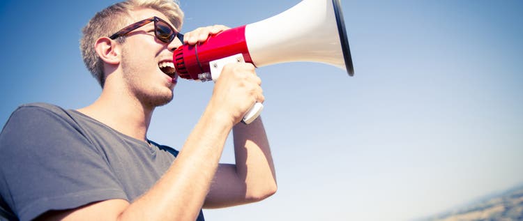 Young caucasian man screaming with a megaphone on the top of a hill during a sunny day.