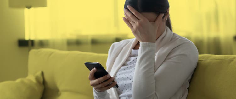 Woman sitting on a couch holding a cellphone and covering her face with one hand