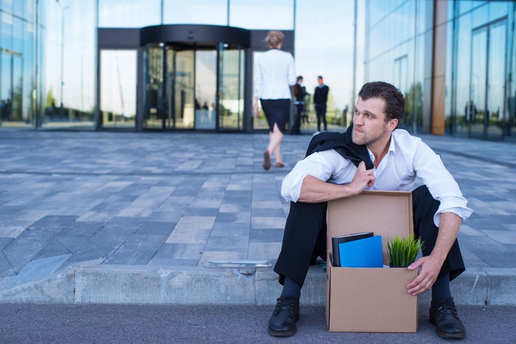 Fired business man sitting frustrated and upset on the street near office building with box of his belongings.