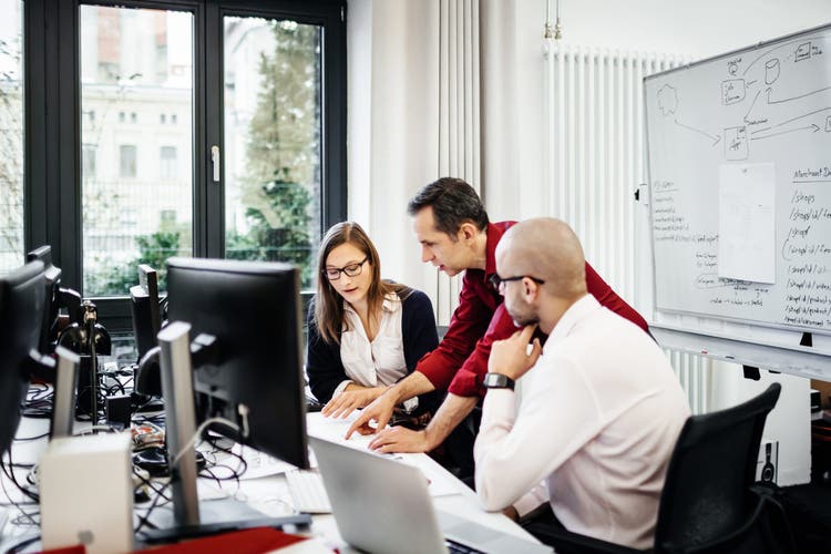 Two of the colleagues are sitting whilst another one is standing and bending over explaining something to them. The office is bright with big windows and a white board is seen with notes on it.