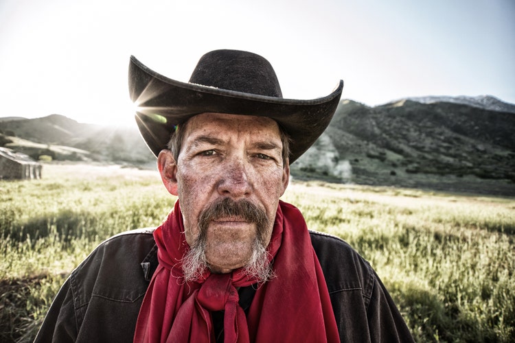 Mature cowboy in a black hat looking like a western outlaw grimacing directly at the camera. Desaturated with added grunge.