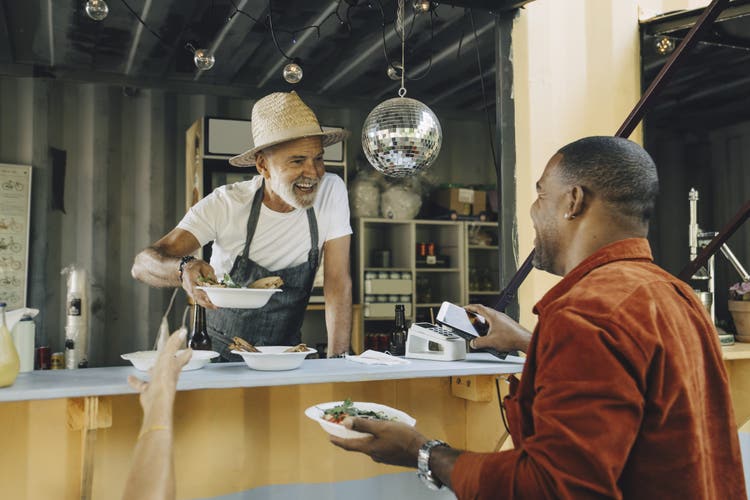 Smiling owner serving food while male customer doing contactless payment through smart phone