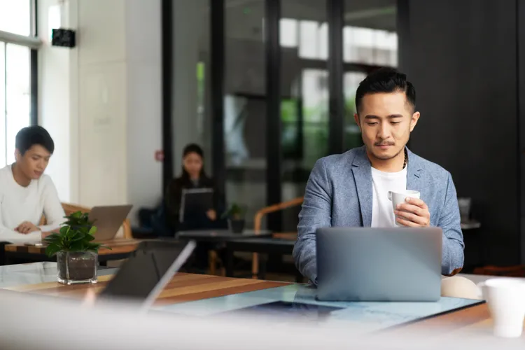 Professional in a blazer holding a coffee while working on a laptop in an office lounge